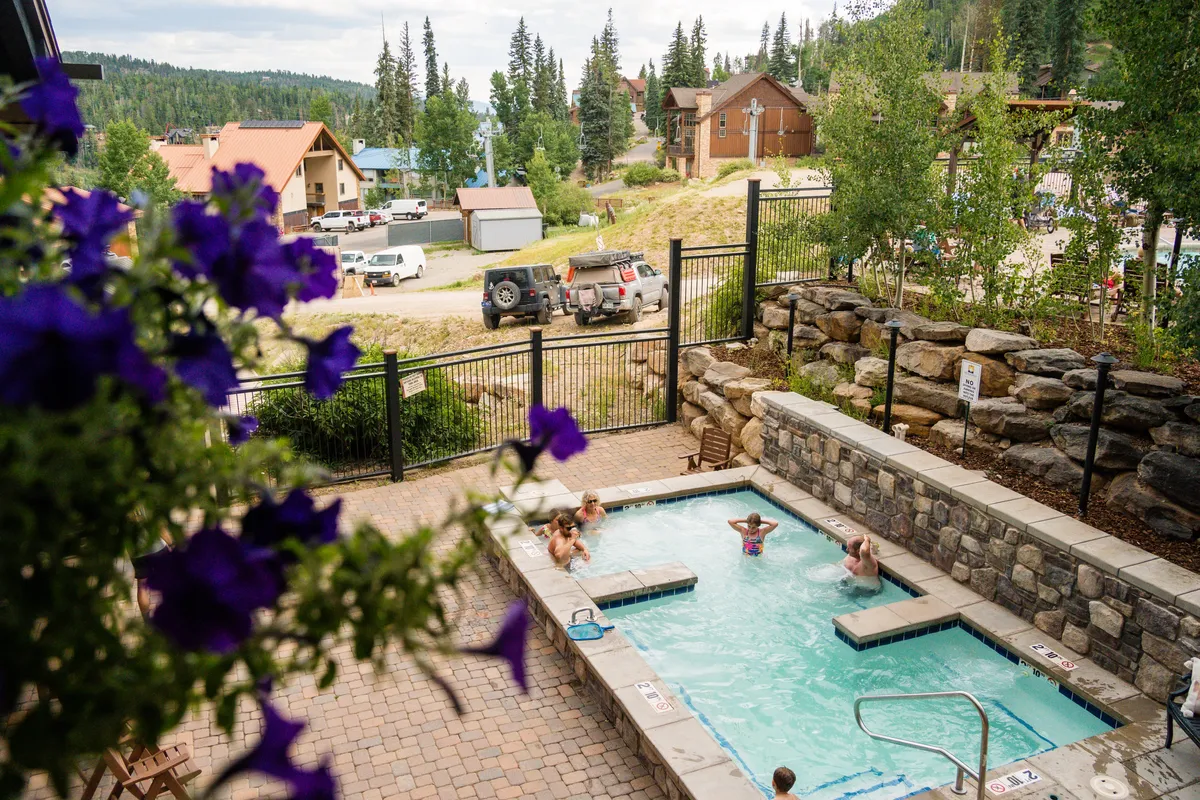 An inviting outdoor hot tub surrounded by greenery, with people relaxing and socializing. The area is landscaped with flowering plants and a rocky wall, creating a lush, resort-like atmosphere.