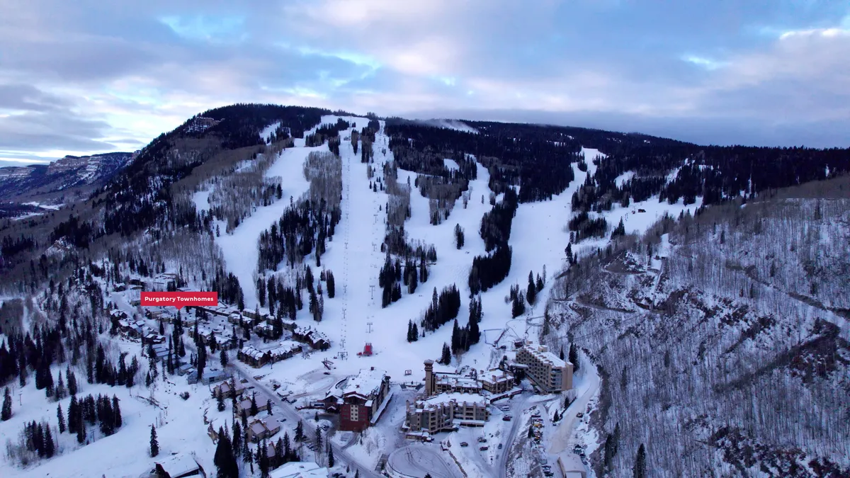 A scenic winter exterior view showcasing the ski slopes, townhouses, mountain backdrop, and the ski lifts. Snow blankets the ground and rooftops, creating a classic ski-in/ski-out atmosphere. The ski lifts are visible in the background, emphasizing direct access to the slopes.