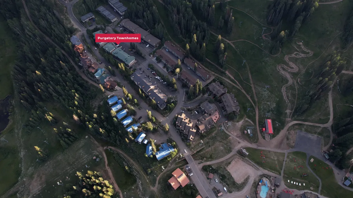 A wide exterior shot of Purgatory Townhomes nestled among tall trees, with ski slopes visible in the background. The setting sun casts a golden glow over the mountains, highlighting both the natural beauty and the proximity to outdoor activities.