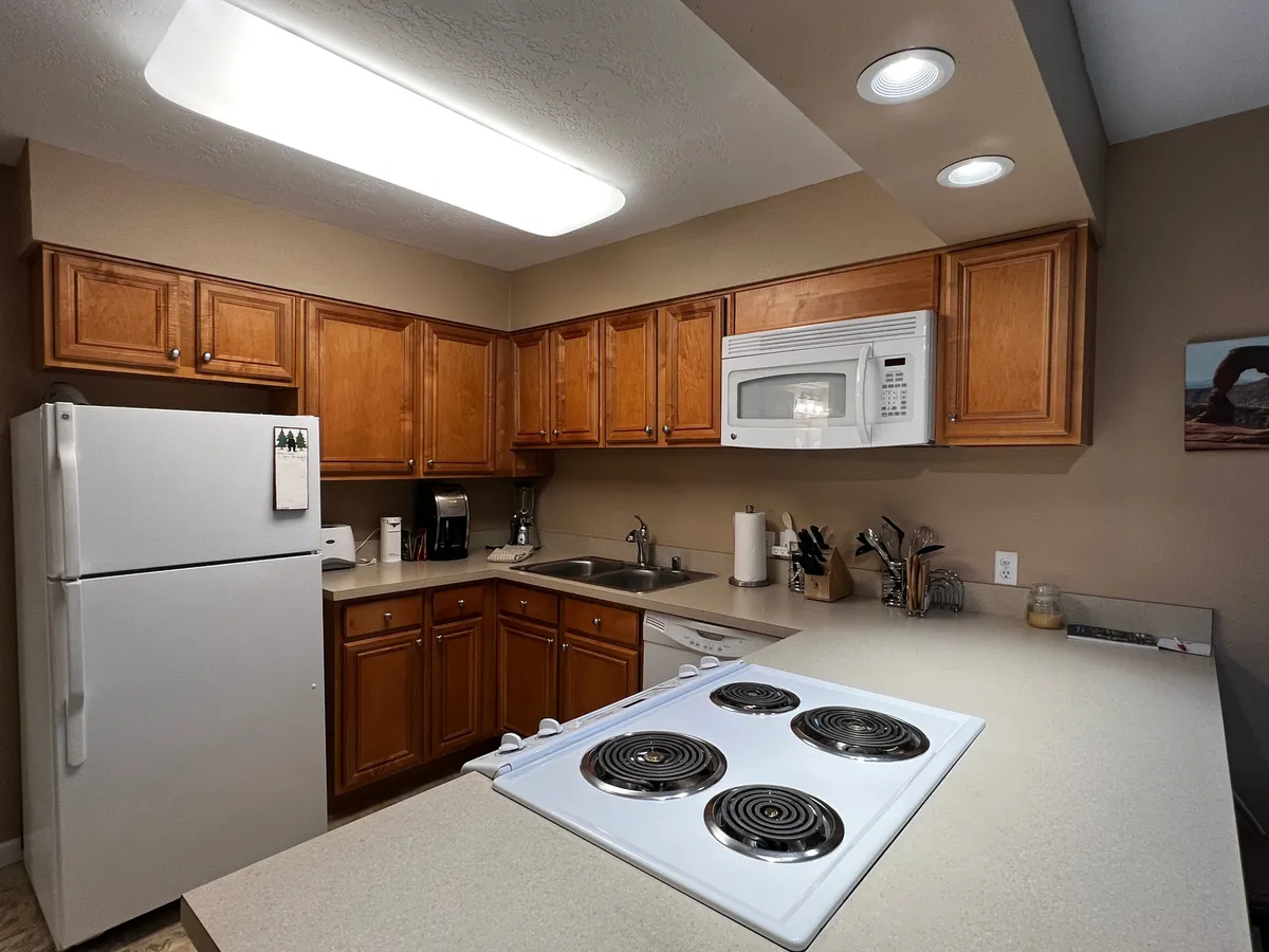 This image features a warm, inviting kitchen with prominent cabinets, a modern stove, a sink, a refrigerator, and expansive countertop space. The scene captures a cozy atmosphere ideal for cooking and entertaining, with hints of family gatherings visible through its organized and accessible layout.