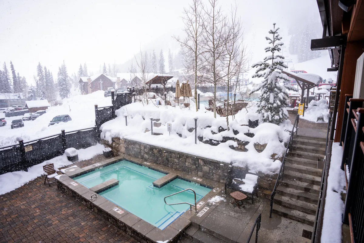 A tranquil exterior scene featuring a steaming hot tub surrounded by snow-covered trees. Steps lead down from the hot tub, suggesting a cozy retreat after a day in the snow. The area feels inviting and peaceful, perfect for après-ski relaxation.