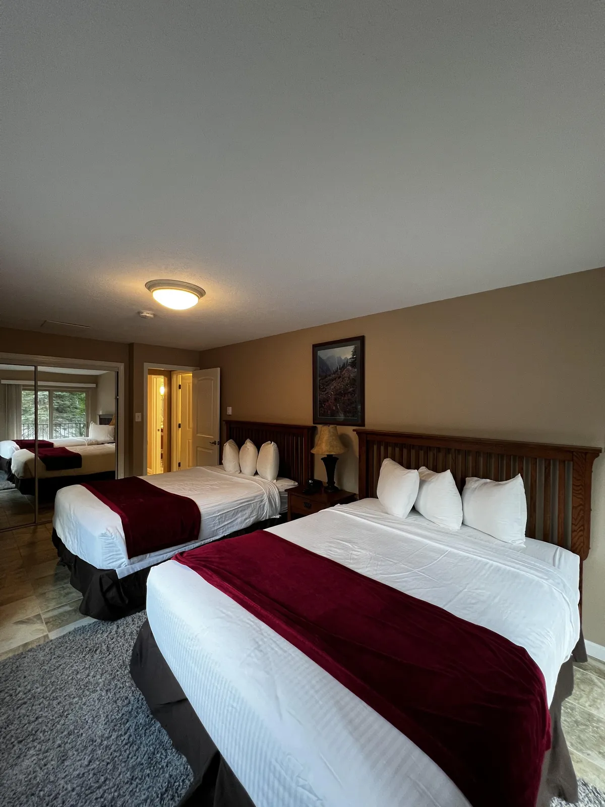 This image features two beds with red throws, a lamp on a bedside table, a framed landscape picture on the wall, and a large window letting in natural light. The space feels airy, inviting, and perfect for relaxing after outdoor adventures or starting the day with views of the landscape.