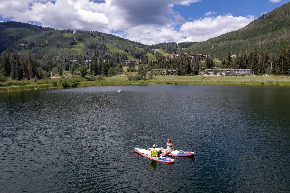 A tranquil lake scene with paddleboarders gliding across the water, mountains rising in the background, and soft clouds reflecting on the lake's surface. The image radiates peace while suggesting adventure and connection to nature.