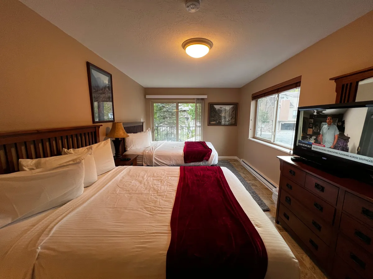 This photo shows a bedroom with two beds adorned with red accent throws, large windows filling the room with natural light, and a television set for entertainment. The room feels spacious and is ideal for relaxing, watching TV, or enjoying the views.