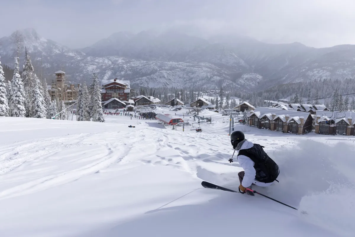 A skier in action on snow-covered slopes with a stunning mountain backdrop and resort buildings visible. The energy and adventure of winter sports are front and center, with the resort's amenities in the vicinity.