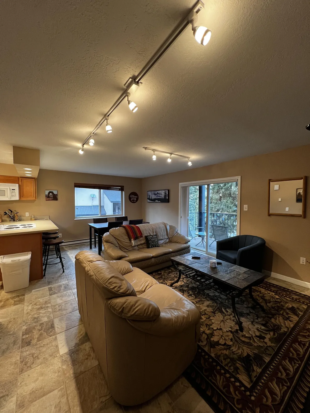 The image showcases a warm, inviting living area featuring plush sofas around a coffee table, an open kitchen in the background, and a sliding glass door leading out to a scenic balcony. Natural light streams in, hinting at mountain views beyond, while the cozy setup invites relaxation and togetherness.